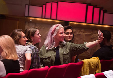 Blonde woman wearing green jacket and pink bandanna surrounded by friends and taking a selfie