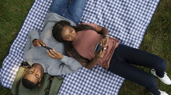 A man and a woman lounge on a picnic blanket playing mobile games.